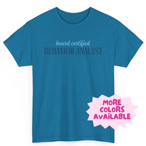 May include: A teal blue t-shirt with the text "board certified BEHAVIOR ANALYST" printed on the front. The text is in a light blue color. A pink starburst sticker with the text "MORE COLORS AVAILABLE" is on the bottom right corner of the shirt.
