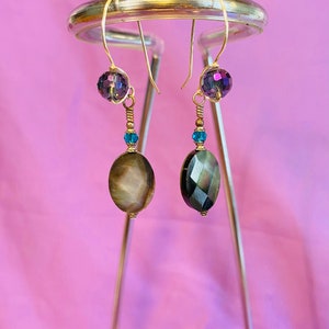 May include: A pair of dangle earrings with a gold wire wrapped around a dark iridescent oval bead, a smaller teal bead, and a dark purple faceted bead. The earrings are hanging from a silver stand.