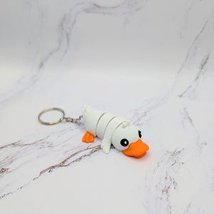 Puede incluir: Llavero de pato de goma blanco con pico y patas naranjas. El pato está enroscado en una posición de dormir.