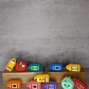 Puede incluir: Una colección de coloridas gomas de borrar con forma de crayones, lápices y manzanas. Las gomas de borrar son de varios colores, incluyendo amarillo, naranja, rosa y azul, con caras sonrientes. Las gomas de borrar se exhiben en un bloque de madera.