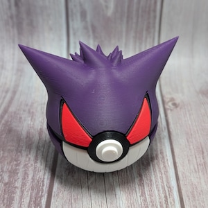 May include: A 3D-printed purple Gengar head atop a white and red Poke Ball. The Gengar has sharp, pointed ears and a spiky head. The Poke Ball has a black band and a white button.