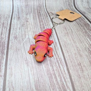 Può includere: Un portachiavi a forma di lucertola rosa, rosso e arancione con un anello portachiavi argentato. La lucertola ha una superficie testurizzata e grandi occhi neri.