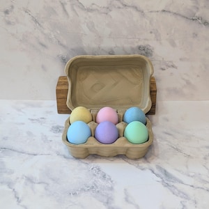 May include: A cardboard egg carton holding six pastel-colored eggs: yellow, pink, blue, purple, and green. The eggs are arranged in two rows of three. The carton is open, revealing the eggs against a marble background.