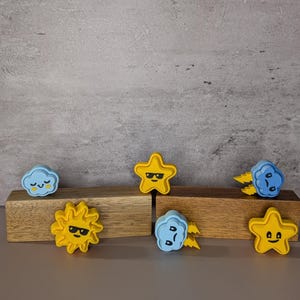 May include: A selection of quirky weather-themed decorations. The set features yellow stars, a yellow sun, and blue clouds, each with a distinct facial expression. The items are displayed on wooden blocks against a grey background.