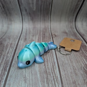 Puede incluir: Un llavero de delfín iridiscente azul y verde con una etiqueta marrón y un llavero plateado. El delfín está hecho de plástico y tiene un cuerpo segmentado. El texto "Ocean Silk" está impreso debajo del delfín.