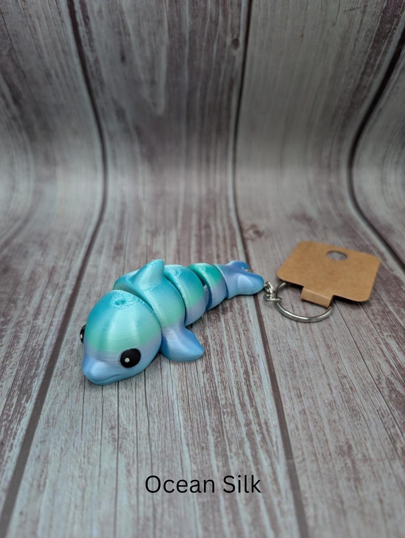 May include: A blue and green iridescent dolphin keychain with a brown tag and a silver key ring. The dolphin is made of plastic and has a segmented body. The text "Ocean Silk" is printed below the dolphin.