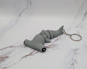 Könnte beinhalten: Ein grauer Hammerhai-Schlüsselanhänger mit einem silbernen Metallring. Der Hai ist aus weichem, biegsamem Material gefertigt und hat ein realistisches Design.