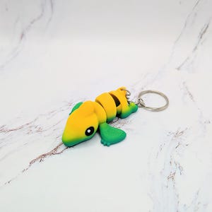 May include: A yellow and green lizard keychain with a silver key ring. The lizard has a segmented body and a cute face.