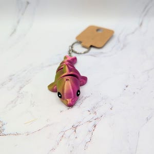 May include: A small, pink and green dolphin keychain made with a 3D printer. The keychain is made with a dual-color filament, creating a gradient effect. The text "Silk Dual - Magenta and Lime" is visible below the dolphin.