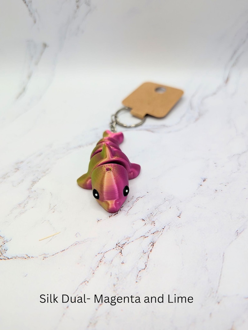 May include: A small, pink and green dolphin keychain made with a 3D printer. The keychain is made with a dual-color filament, creating a gradient effect. The text "Silk Dual - Magenta and Lime" is visible below the dolphin.