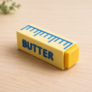 May include: A yellow butter dish with blue text that reads "BUTTER" and a blue ruler design on top. The dish has a small, removable yellow lid. The butter dish is on a light brown wooden surface.
