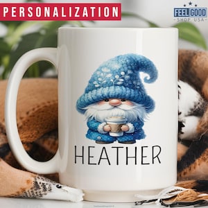 May include: A white ceramic coffee mug with a blue gnome illustration holding a cup of coffee. The gnome is wearing a blue knit hat with white snowflakes. The text 'HEATHER' is printed below the gnome.
