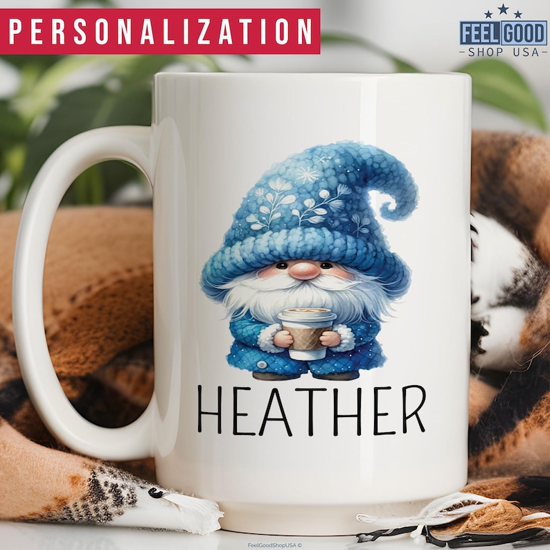 May include: A white ceramic coffee mug with a blue gnome illustration holding a cup of coffee. The gnome is wearing a blue knit hat with white snowflakes. The text 'HEATHER' is printed below the gnome.