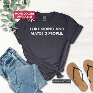 May include: Dark heather gray t-shirt with the text "I LIKE SKIING AND MAYBE 3 PEOPLE." The shirt is styled with blue jeans and brown flip-flops. The words "MORE COLORS AVAILABLE!" are in a pink box.