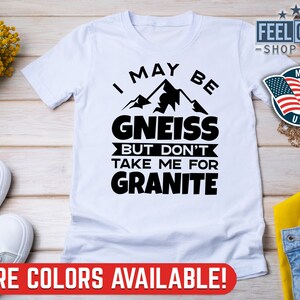 May include: White t-shirt with a black graphic that says "I may be gneiss but don't take me for granite". The graphic also includes a mountain range. The shirt has a v-neck and short sleeves.