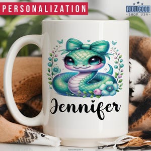 May include: White ceramic mug featuring a green snake with a pink bow and flowers. The mug is personalised with the name "Jennifer".