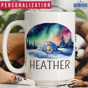 May include: White ceramic mug with a watercolor illustration of a snow-covered landscape with a small igloo and a colorful aurora borealis in the sky. The text "HEATHER" is printed on the mug.