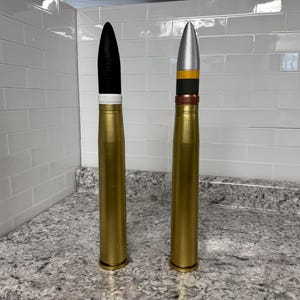 May include: Two large, gold-coloured artillery shells with different coloured tips. The shell on the left has a black tip with a white band, while the shell on the right has a silver tip with a black band and a yellow band.