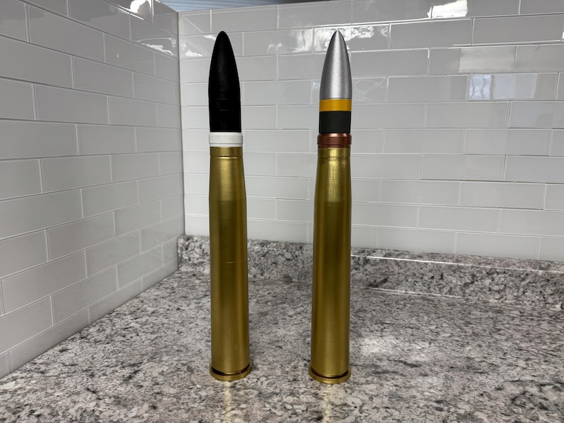 May include: Two large, gold-coloured artillery shells with different coloured tips. The shell on the left has a black tip with a white band, while the shell on the right has a silver tip with a black band and a yellow band.