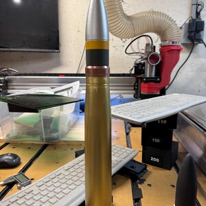 May include: A gold-coloured artillery shell with a silver tip and black and yellow bands. The shell is sitting on a wooden surface with a keyboard and other objects in the background.