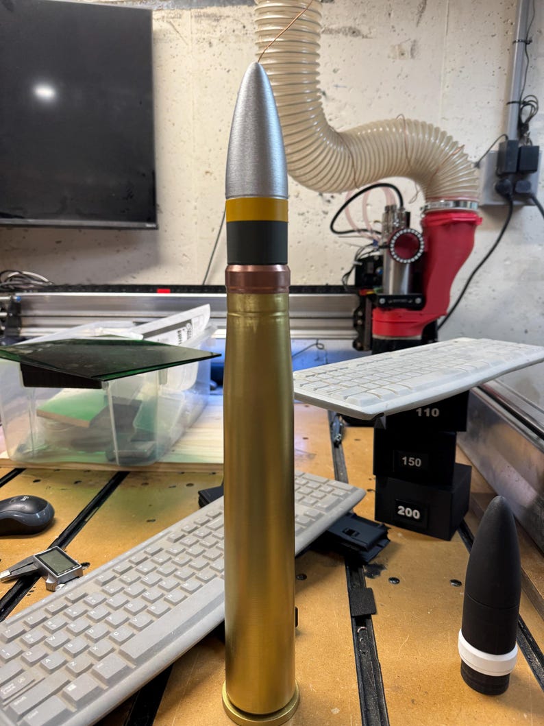 May include: A gold-coloured artillery shell with a silver tip and black and yellow bands. The shell is sitting on a wooden surface with a keyboard and other objects in the background.