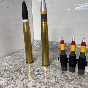May include: Four gold-coloured artillery shells with black, red and yellow painted bands. The shells are arranged in a row on a grey countertop. Two of the shells are larger than the other two.