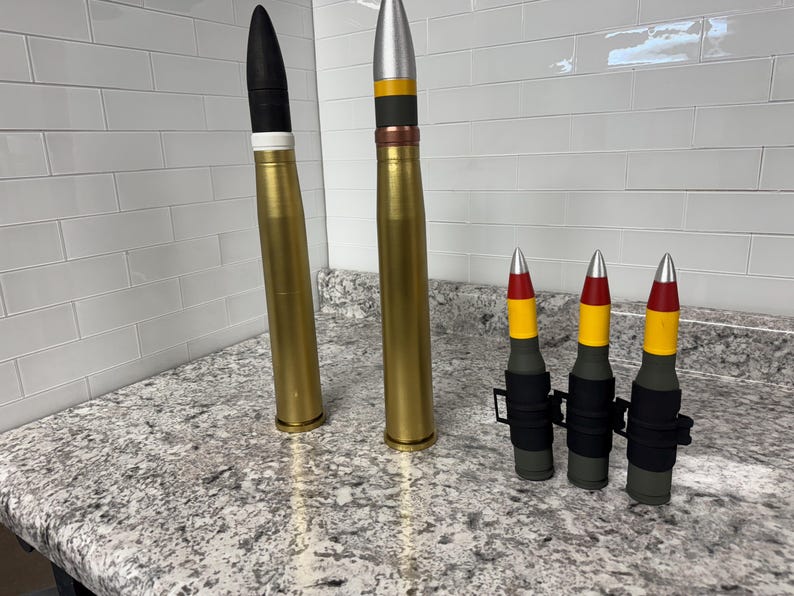 May include: Four gold-coloured artillery shells with black, red and yellow painted bands. The shells are arranged in a row on a grey countertop. Two of the shells are larger than the other two.