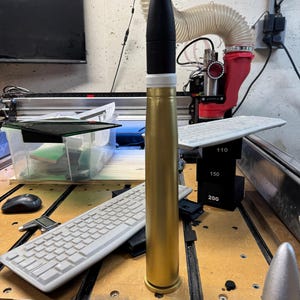 May include: A gold-coloured artillery shell with a black tip and a white band. The shell is standing upright on a wooden surface. There is a keyboard, a pair of pliers, and a ruler in the background.