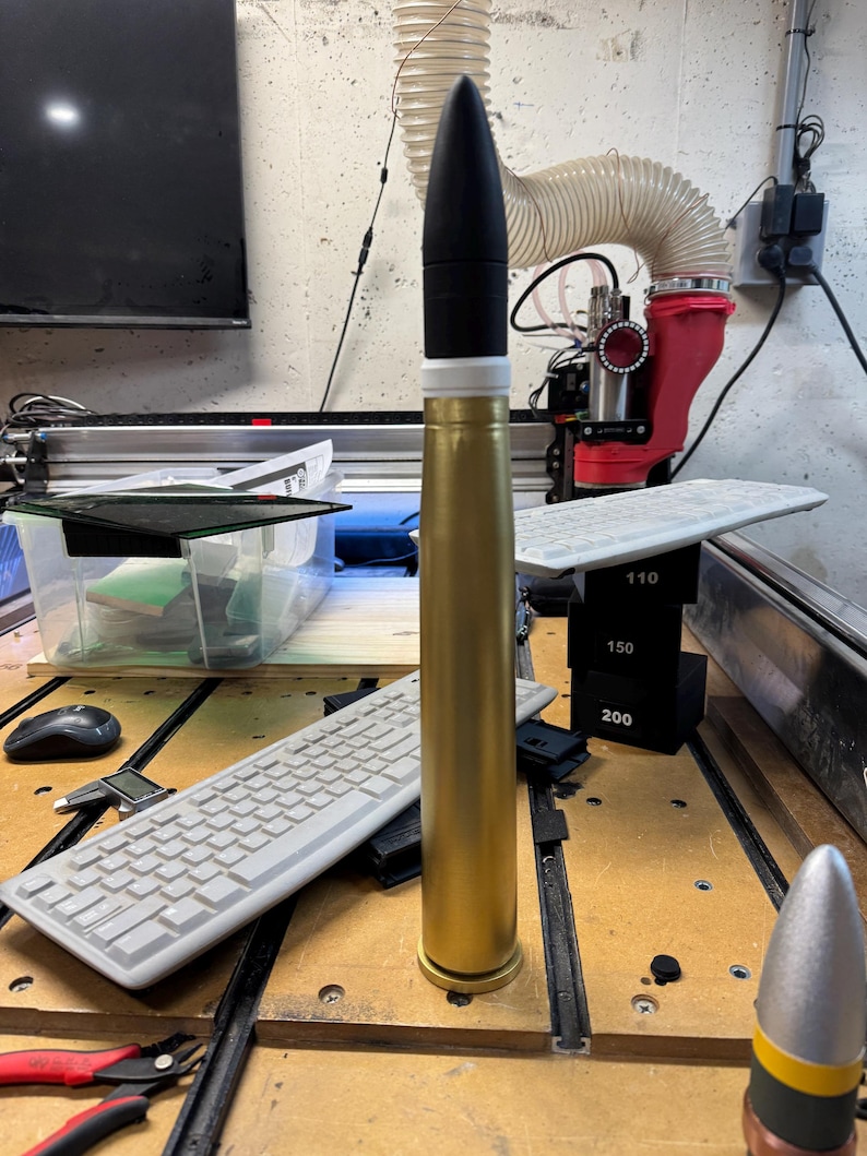 May include: A gold-coloured artillery shell with a black tip and a white band. The shell is standing upright on a wooden surface. There is a keyboard, a pair of pliers, and a ruler in the background.