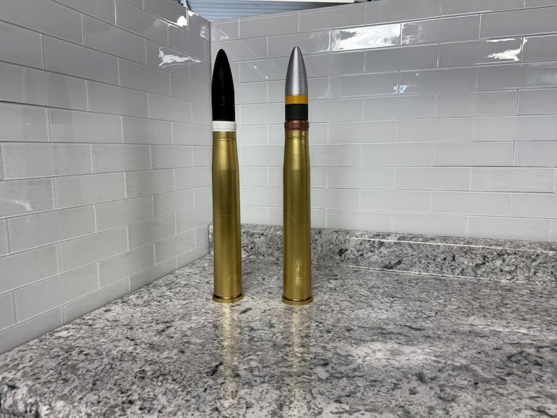 May include: Two gold-coloured metal artillery shells, one with a black tip and white band, the other with a silver tip and black, yellow and red bands.