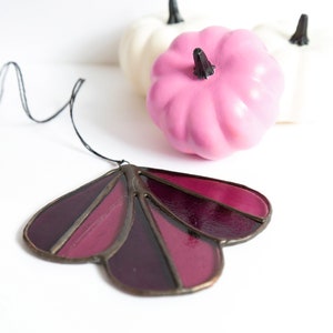 May include: A stained glass suncatcher in the shape of a flower with five petals. The petals are shades of burgundy and purple. The suncatcher is hanging from a black string.