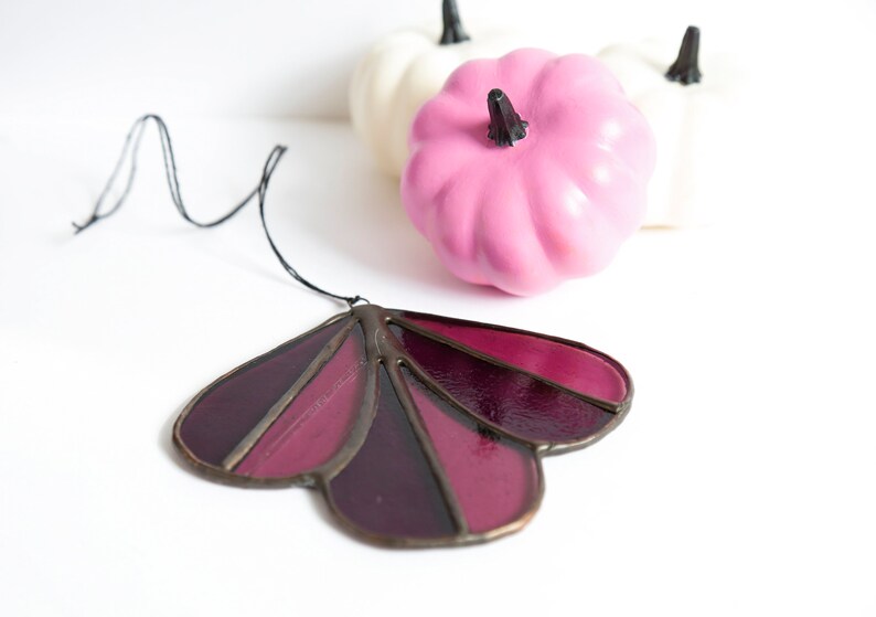 May include: A stained glass suncatcher in the shape of a flower with five petals. The petals are shades of burgundy and purple. The suncatcher is hanging from a black string.