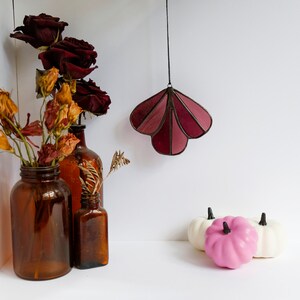 May include: Three amber glass bottles with dried flowers and a stained glass hanging ornament in the shape of a shell. The ornament is burgundy and pink. Three small white pumpkins and one pink pumpkin are on a white surface.