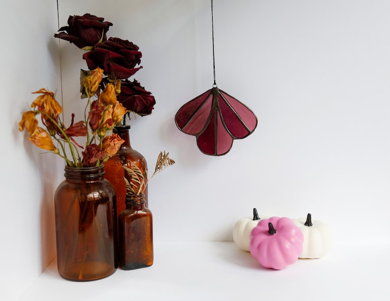 May include: Three amber glass bottles with dried flowers and a stained glass hanging ornament in the shape of a shell. The ornament is burgundy and pink. Three small white pumpkins and one pink pumpkin are on a white surface.