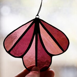 May include: A stained glass ornament in the shape of a flower with five petals. The petals are made of pink and red glass and are outlined in black. The ornament is hanging from a black string.
