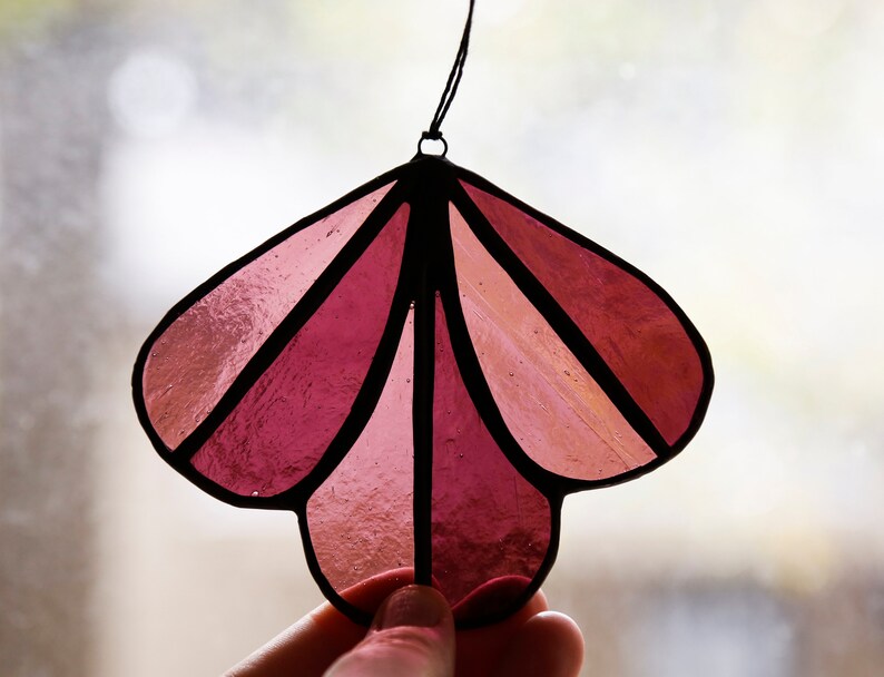 May include: A stained glass ornament in the shape of a flower with five petals. The petals are made of pink and red glass and are outlined in black. The ornament is hanging from a black string.