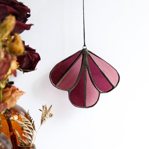 May include: A stained glass suncatcher in the shape of a flower with a pink and purple gradient. The suncatcher is hanging from a black string.