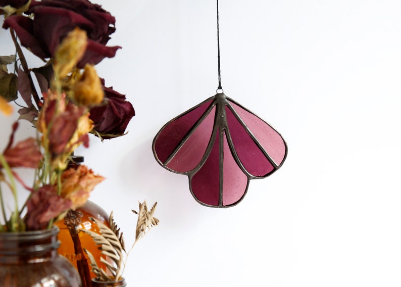 May include: A stained glass suncatcher in the shape of a flower with a pink and purple gradient. The suncatcher is hanging from a black string.