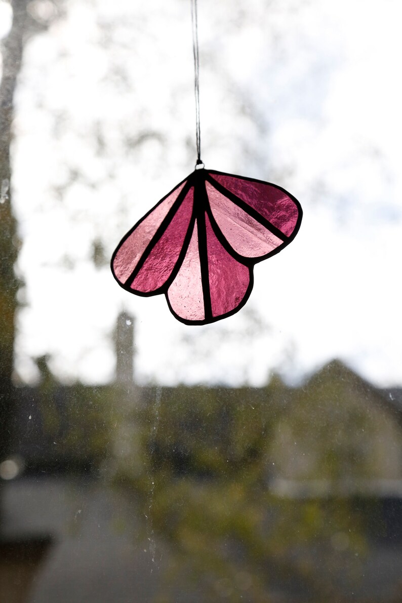 May include: A pink stained glass suncatcher shaped like a flower with five petals. The suncatcher is hanging from a string.