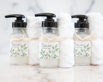 May include: Three clear glass bottles with black pump dispensers, filled with a light liquid. Each bottle has a label with the text "The Smiths July 19th, 2025" and a green leaf design. Small white towels are wrapped around the bottles, tied with cream-colored ribbons.