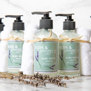 May include: A set of six clear glass bottles with pumps, each filled with a clear liquid. The bottles are labeled with a green and white label that reads "SUDS & snuggles BABY SMITH ARRIVING November 2023". Each bottle is wrapped with a white washcloth and a beige ribbon. The bottles are arranged on a white marble surface with dried lavender sprigs.