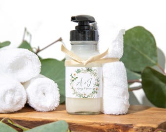 May include: A clear glass bottle with a black pump dispenser filled with a white liquid. The bottle has a white label with green leaves and white flowers. The label says "A + J Always & Forever". The bottle is sitting on a wooden surface with white towels and green leaves.