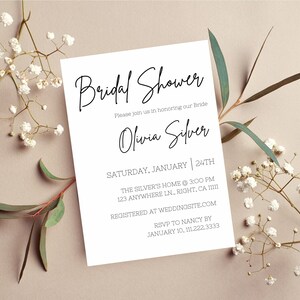May include: A white invitation card with black script that reads "Bridal Shower" and details for the event, including the bride's name, date, time, and location. The card is surrounded by sprigs of white flowers and greenery.