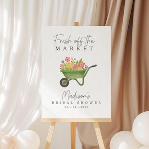 May include: A white sign on a wooden easel with the text "Fresh off the MARKET" above a watercolor illustration of a green wheelbarrow filled with flowers. Below, it reads "Madison's BRIDAL SHOWER 06 • 14 • 2026". White and beige balloons are in the foreground.