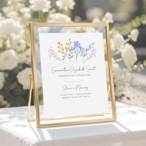 May include: A gold metal frame holding a white sign with a watercolor floral illustration in yellow, blue, and pink. The text reads: Samantha Elizabeth Smith, February 24, 1960 - October 3, 2020, Share a Memory, Please take a card & share a memory with the family.