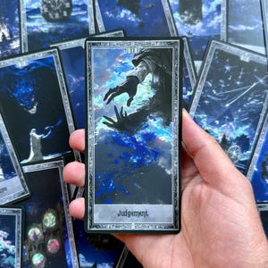 Puede incluir: Una carta del tarot con una ilustración oscura de manos extendidas, con la palabra "Juicio" en la parte inferior. La carta se sostiene en una mano, rodeada de otras cartas del tarot con obras de arte similares.