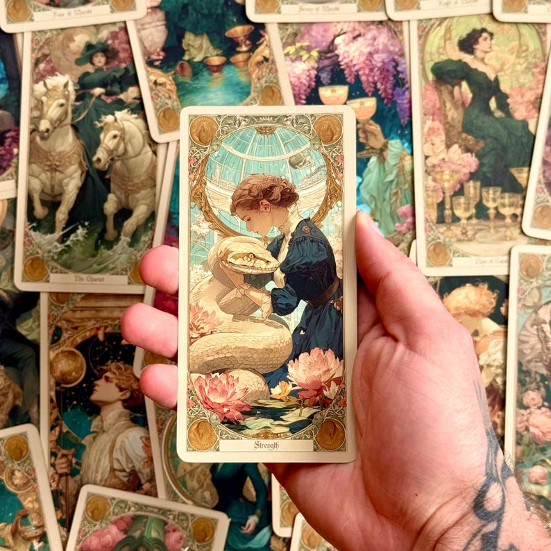Op de afbeelding: Een rechthoekige tarotkaart met de titel Strength, voorzien van een illustratie van een persoon in een donkerblauwe jurk die interactie heeft met een grote witte slang. Het tafereel is omlijst door een gouden rand met bloemmotieven.