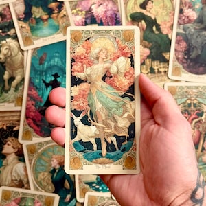 Op de afbeelding: Een rechthoekige tarotkaart getiteld The World met een afbeelding van een figuur in een zwierige jurk omringd door roze bloemen en een wit hert, tegen een donkerblauwe sterrenhemel met een gouden rand en cirkelvormige accenten.