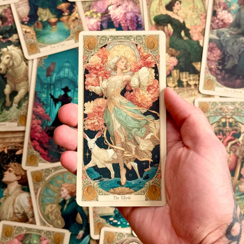 Op de afbeelding: Een rechthoekige tarotkaart getiteld The World met een afbeelding van een figuur in een zwierige jurk omringd door roze bloemen en een wit hert, tegen een donkerblauwe sterrenhemel met een gouden rand en cirkelvormige accenten.