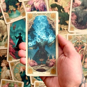 Op de afbeelding: Een rechthoekige tarotkaart getiteld The Mage met een figuur met een hoofd van blauwe rook die een gloeiende bol vasthoudt. De sc&egrave;ne bevat een tafel met een kelk, een glazen fles, kaarsen en roze bloemen, omlijst door een sierlijke gouden rand.